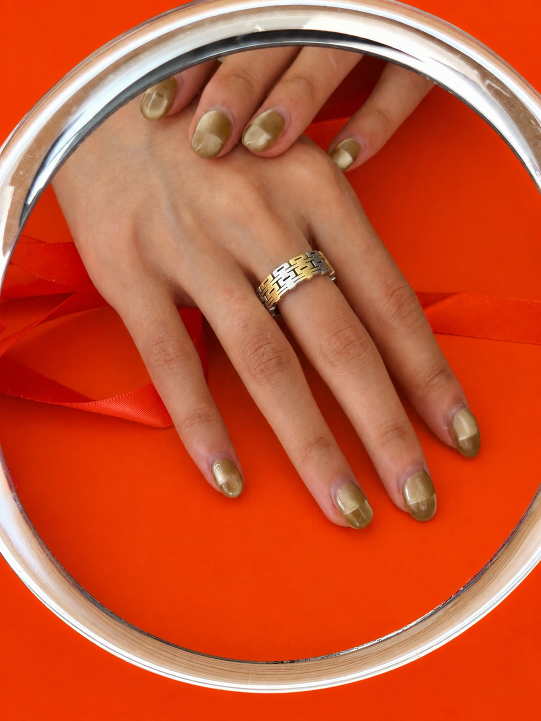 A close-up of a hand wearing a two-tone modern Chinese-inspired infinite happiness symbol ring reflected in a round mirror. The background is a vibrant orange with a matching orange ribbon.