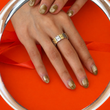 A close-up of a hand wearing a two-tone modern Chinese-inspired infinite happiness symbol ring reflected in a round mirror. The background is a vibrant orange with a matching orange ribbon.