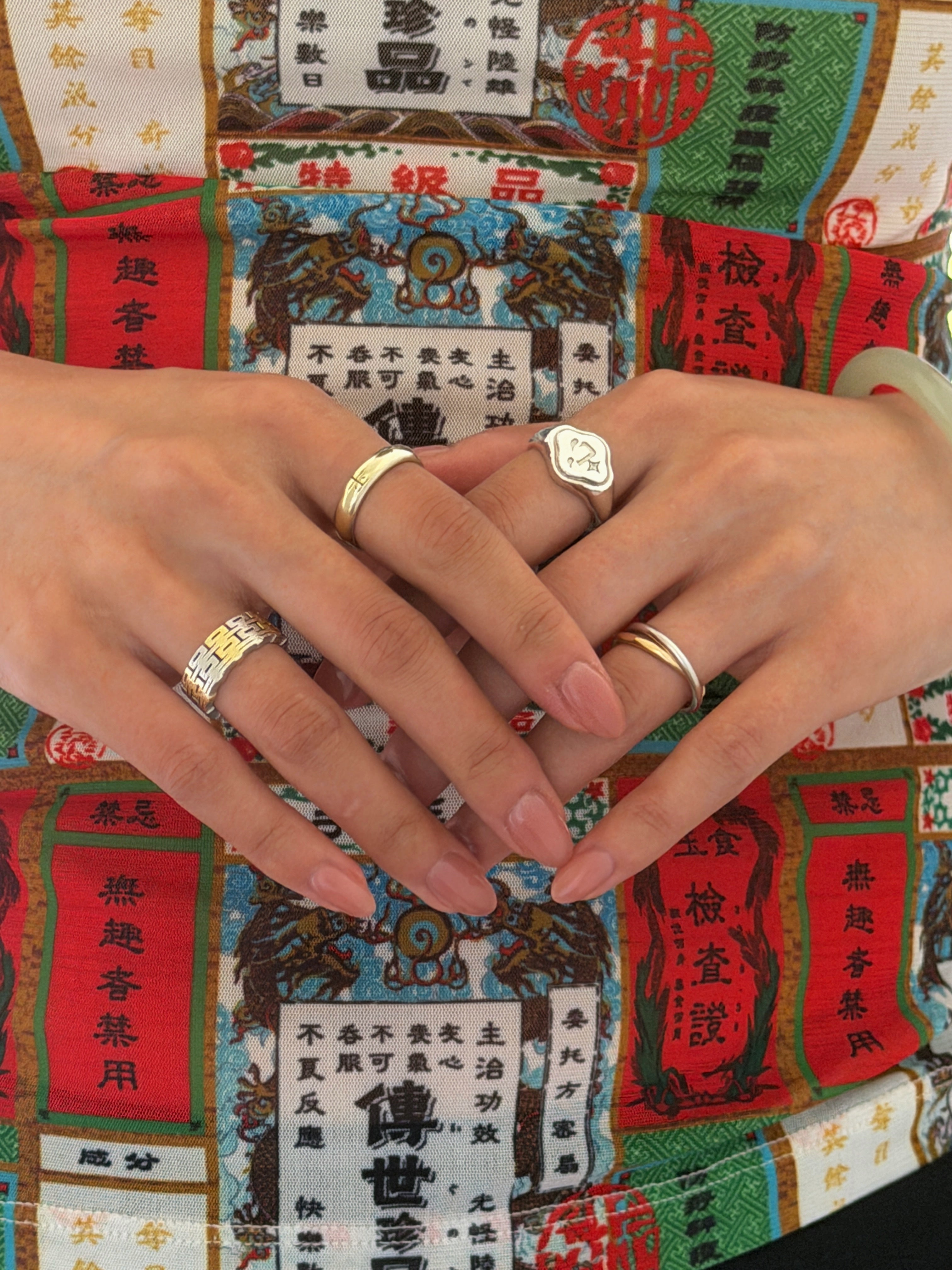 A close-up of two hands wearing several modern Chinese-inspired rings that are in gold, silver, and two-tone.